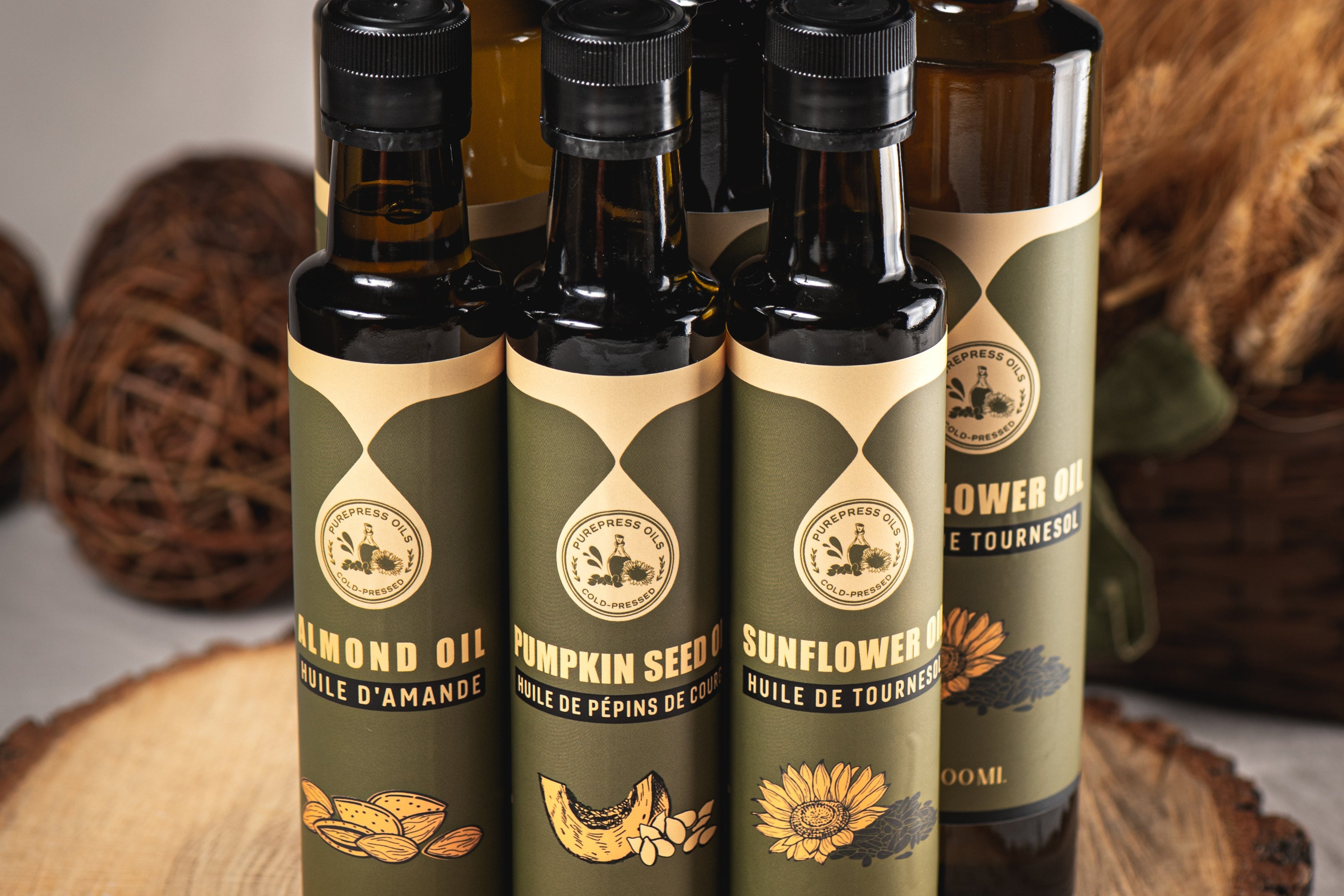 Six bottles of cold-pressed oils, including almond, pumpkin seed, and sunflower oil, displayed on a rustic wooden slab with seeds scattered in front. The background features natural elements like wheat and wicker, creating a warm, artisanal feel.